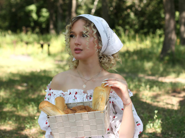 AnfisaRalphs with curly blonde hair, white headscarf, off-shoulder floral dress, holding basket of baguettes, sunny forest, dappled light, gold necklace, soft expression, attractive, tits visible.