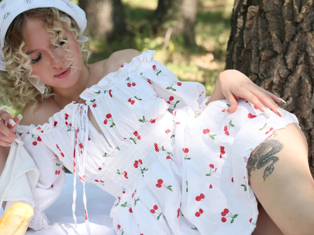 AnfisaRalphs with curly blonde hair, wearing a white cherry dress, lying against a tree, showing a thigh tattoo, and holding a baguette. Sunlight highlights her dress and skin.