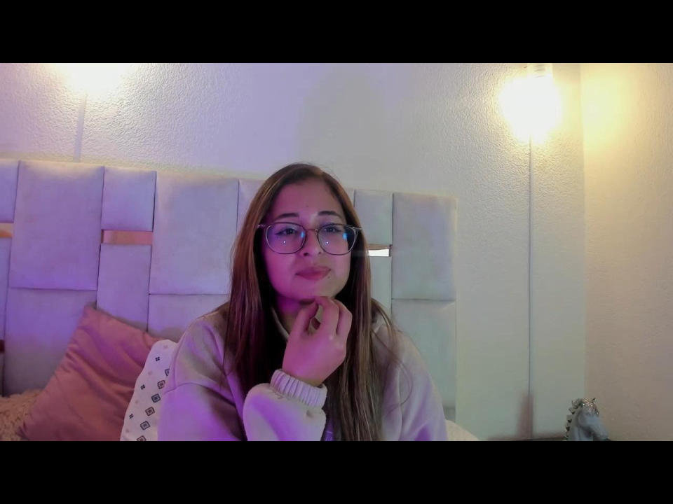 SophiaCollinsX with glasses, long brown hair, pink sweater, hand under chin, smiling, soft pink and white headboard, warm light, cozy bedroom setting, purple and blue lighting.