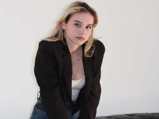 EmelyHailey met blond haar, een zwart blazer, een wit tanktop en blauwe jeans, leunt voorover, waarbij haar kleine borsten zichtbaar zijn, en draagt een ketting met een ster aan..