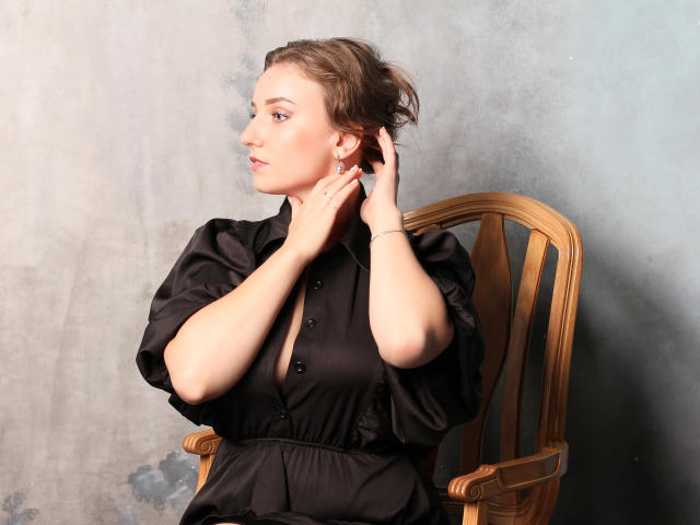 Kristi Watson, con i capelli castani corti, indossa un abito nero con bottoni, si tocca l'orecchio con entrambe le mani, è seduta su una sedia di legno e guarda di lato con un leggero sorriso..