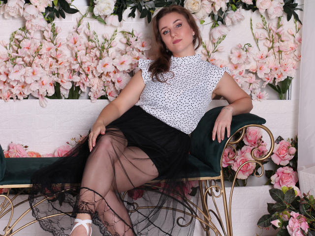 Kristi Watson, con i capelli castani ricci, indossa una camicetta bianca a pois, una gonna nera e collant neri trasparenti, seduta su una poltrona verde in velluto, con le gambe incrociate, mostrando il sedere..