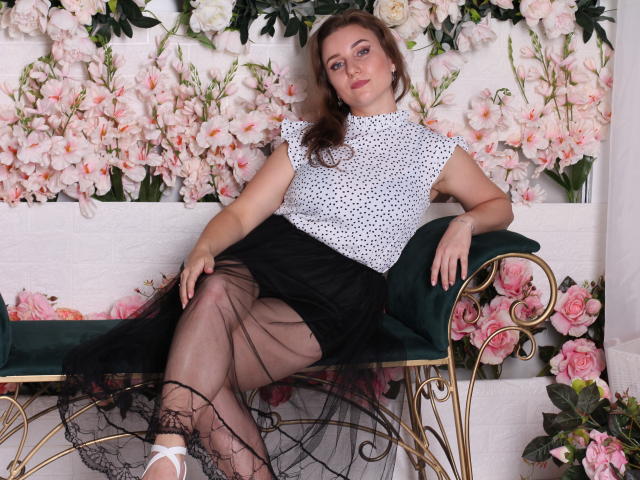 Kristi Watson, con i capelli castani, una camicetta bianca a pois, una gonna nera trasparente e collant neri, seduta su una panca di velluto verde, mostra i seni e i glutei, circondata da fiori rosa..