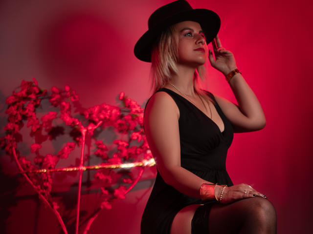 PerlaDivaio with blonde hair, black hat, black dress, garter, and thigh-high stockings, red lighting, holding hat, looking to side, gold bracelet, sexy, curvy, medium tits.