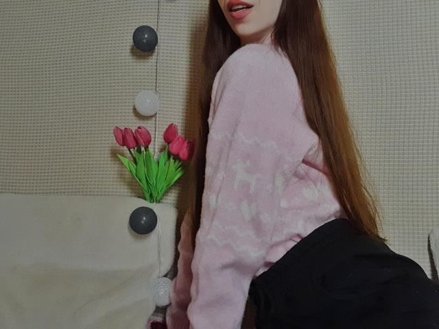 LourdesRay with long brown hair, pink fuzzy sweater with white patterns, black skirt, and pink tulips in her mouth's right. She's kneeling, showing her ass, on a beige couch.