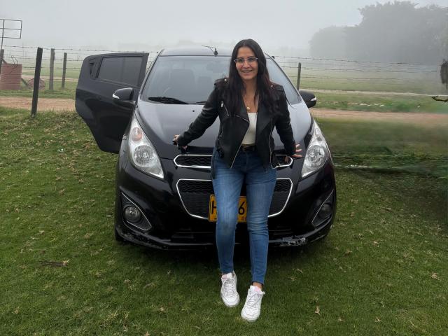 AnnaRous è in piedi di fronte a una macchina nera con la portiera aperta, indossa una giacca di pelle, una maglietta bianca, jeans blu e scarpe da ginnastica bianche, con lunghi capelli scuri..