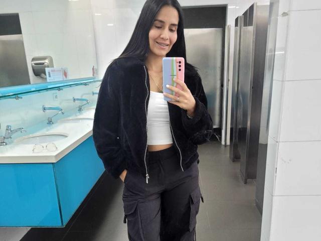 AnnaRous hat langes, schwarzes Haar, ein süßes Lächeln und trägt eine schwarze Jacke, ein weißes Crop-Top und schwarze Hosen. Sie macht ein Selfie vor einem Spiegel in einem Badezimmer mit blauen Waschbecken..