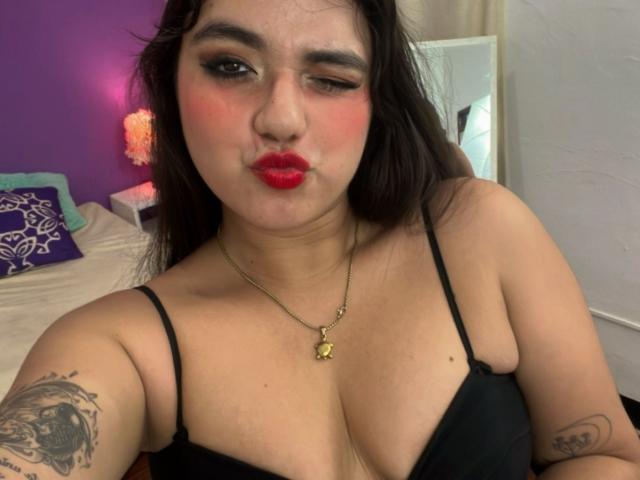 VictoriaSaens, with winking eye and red lips, wearing black bra, showing her big tits and tattooed arm, wearing gold necklace with star pendant, looking cute.