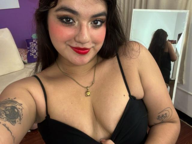 VictoriaSaens con rossetto rosso, reggiseno nero, che mostra un seno abbondante, tatuaggi sulle braccia, collana d'oro, riflesso nello specchio, capelli scuri, pelle chiara, sorridente, che si fa un selfie in camera da letto..