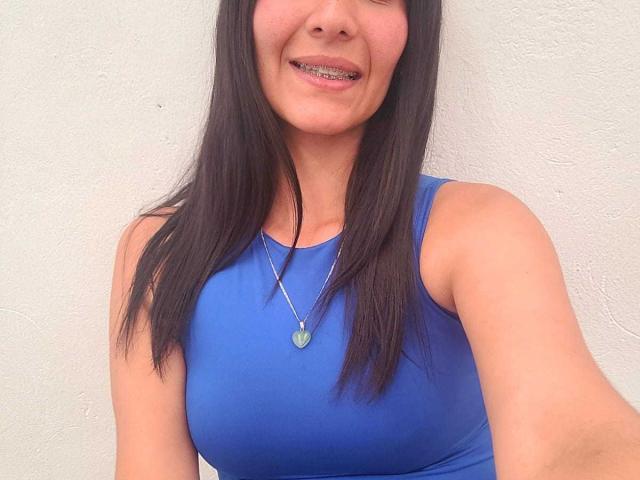 AnnaRous has long black hair, a blue sleeveless top, and a silver necklace with a green pendant. She's smiling with her teeth showing. Boobs are visible.