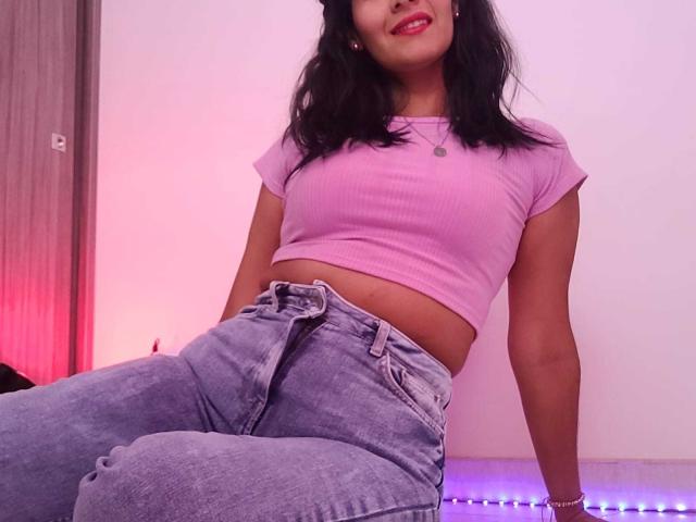 AnnaRous, con i capelli neri, indossa un top corto a coste rosa che lascia scoperto il ventre, e jeans a vita alta, di colore azzurro chiaro, seduta con un leggero sorriso, con una luce viola sullo sfondo..