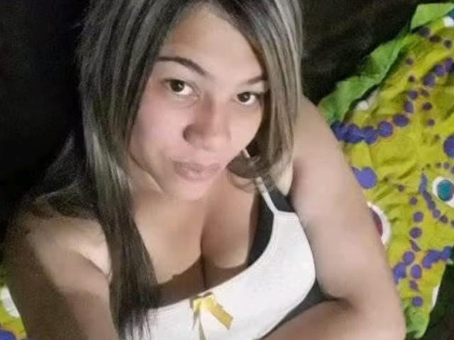 AlmaWhite69 with gray hair, brown eyes, and a pouty face, wearing a white tank top showing her large, rounded tits. She's sitting on a colorful green and purple pillow.