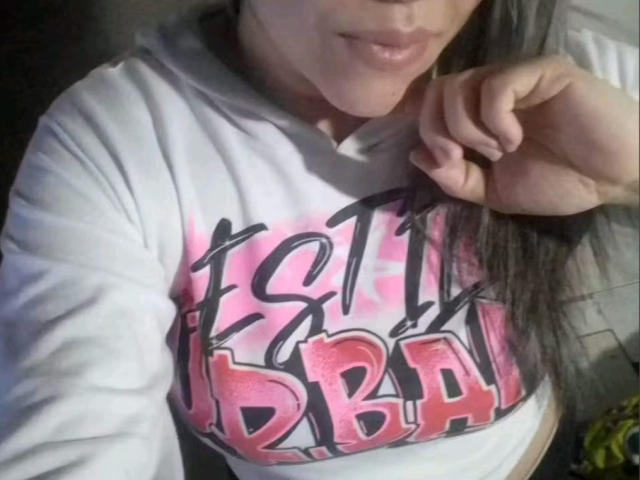 AlmaWhite69 con lunghi capelli scuri, indossa una felpa bianca, mostrando i suoi piccoli seni e le labbra leggermente dischiuse, tiene il bordo della felpa, testo in stile graffiti rosa e nero..