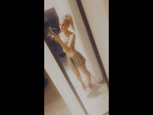 LianaLou69-hot with blonde ponytail, small breasts, wearing leopard print crop top and skirt, taking mirror selfie, holding phone, standing on white floor, in dimly lit room.