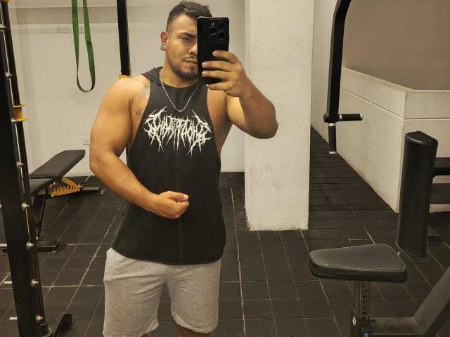 BrianMarly, muscular, tan-skinned, short black hair, black tank top with white graffiti, gray shorts, holding phone, mirror selfie, gym, standing, right arm flexed, black tile floor.