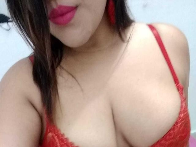 PaulinaQueens with red lace bra, showing her big boobs, red lipstick, and brown hair, wearing red earrings, in a selfie.