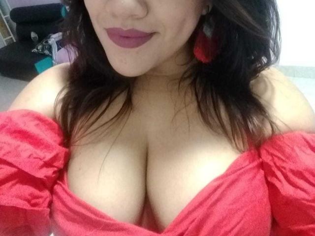 PaulinaQueens with dark hair, red lipstick, and red off-shoulder top, showing her large, cleavage-filled tits. She's wearing red earrings, smiling slightly, and has a cute nose.