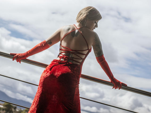 KatParadise con un vestido rojo de terciopelo con espalda con cordones, guantes rojos y cabello rubio corto, mostrando tatuajes, sosteniendo una barandilla, con su trasero en el centro..