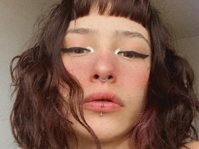 PinkyPieX has wavy brown hair, a nose ring, and a lip piercing with a small ball. She's wearing bold black eyeliner with white winged accents.