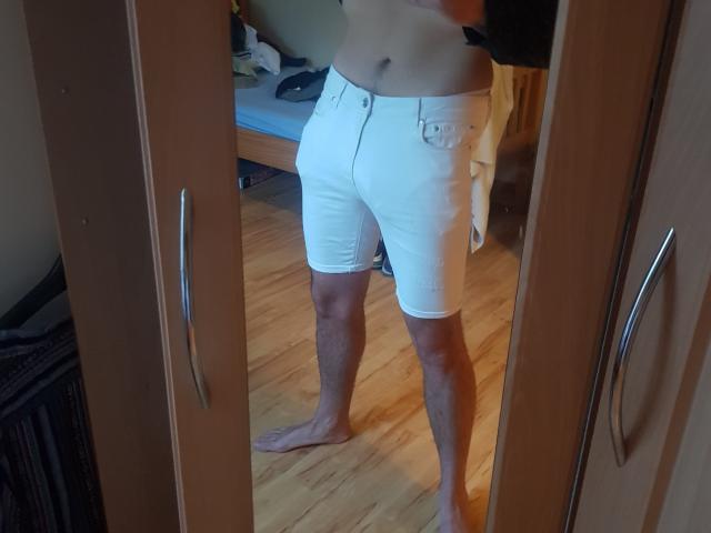 PrettyCharmer's bare torso and white shorts with a button and pocket, standing barefoot on a wooden floor, reflected in a mirror. Their skin is tan, and they're slightly muscular.