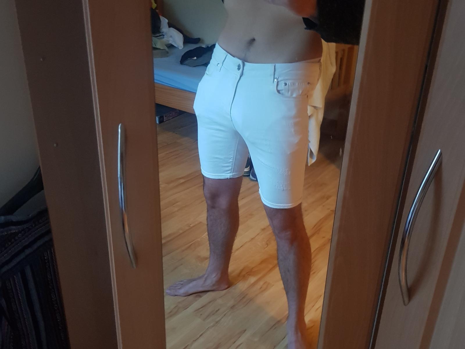 PrettyCharmer's bare torso and white shorts with a button and pocket, standing barefoot on a wooden floor, reflected in a mirror. Their skin is tan, and they're slightly muscular.