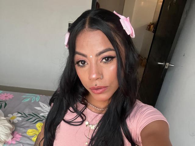 Amberv, avec de longs cheveux noirs, des accessoires pour cheveux en forme de nœud rose, un anneau de nez, un point sur le visage et un collier de perles, portant une chemise rose, regardant l'appareil photo, avec un lit à motifs floraux en arrière-plan..