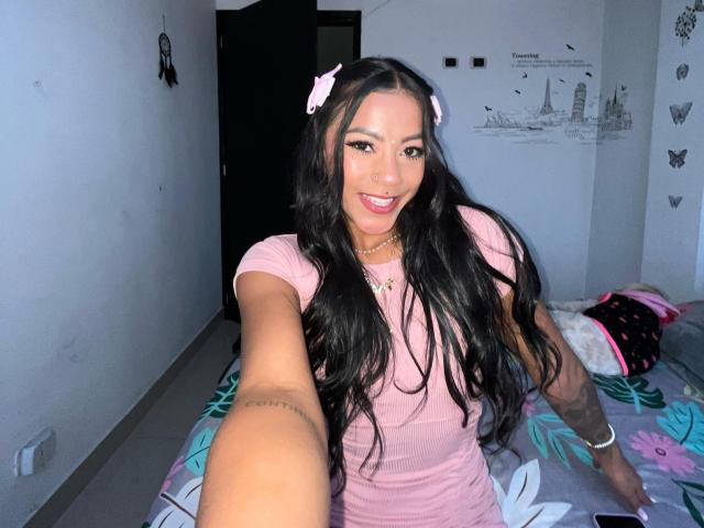 Amberv, con lunghi capelli neri e mollette rosa, indossa una maglietta rosa, sorride alla fotocamera, mostrando un tatuaggio sul braccio destro, e si trova su un letto con lenzuola a fiori..