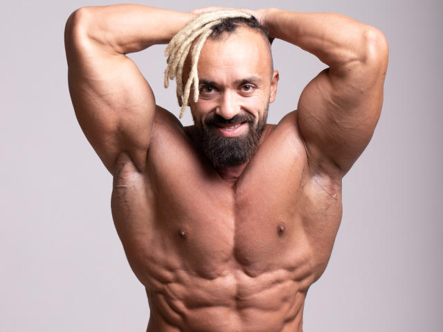 Alexhunk69, muscular, shirtless, bearded, with white dreadlocks, arms raised, showing defined abs and pecs, smiling confidently, gray background.