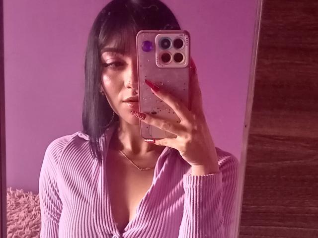 LindaWatsong with black bob, purple ribbed shirt, gold necklace, red nails, holding phone, taking mirror selfie, pink room, wood door, subtle light, focused expression.