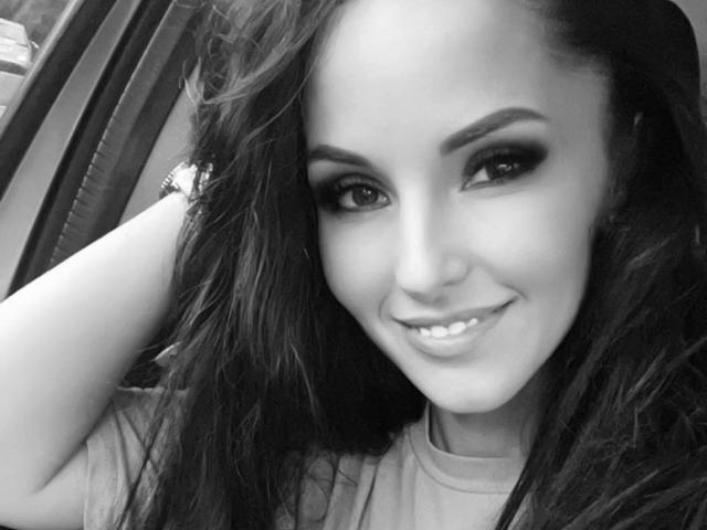 Black-and-white selfie of SweetCarlaHot69 with long dark hair, thick eyebrows, smoky eye makeup, and a cute smile, showing her teeth. She's wearing a shirt and a bracelet.