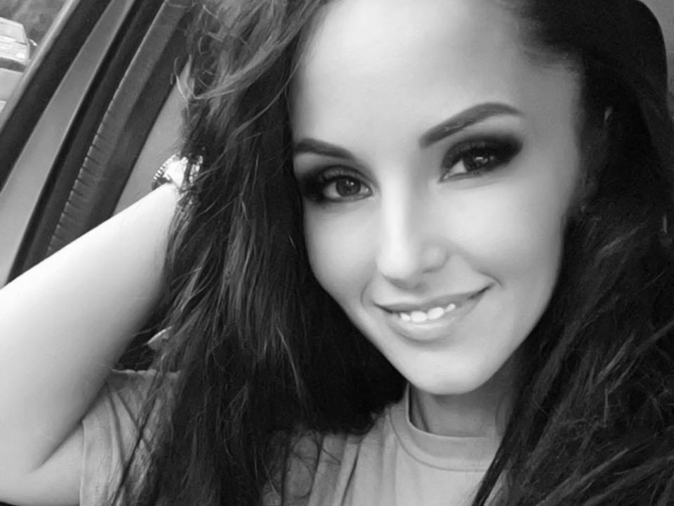 Ein Schwarz-Weiß-Selfie von SweetCarlaHot69 mit langen, dunklen Haaren, dicken Augenbrauen, Smokey-Eye-Make-up und einem süßen Lächeln, bei dem ihre Zähne zu sehen sind. Sie trägt ein Hemd und ein Armband..