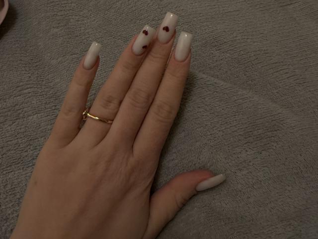 La mano de SugarrX, con uñas largas, blancas y en forma de almendra, un anillo de oro y pequeños puntos rojos en las uñas, descansando sobre una superficie gris texturizada..