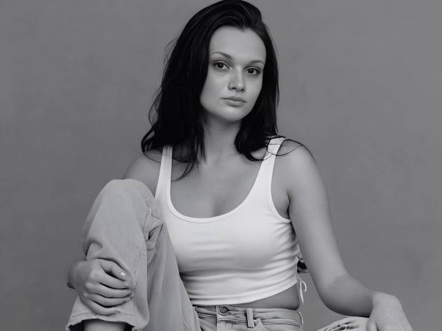 CamilleXMyr, con cabello oscuro, vistiendo una camiseta blanca, mostrando pequeños senos, sentada con las piernas cruzadas, sosteniendo la rodilla, usando jeans, fondo gris, mirando ligeramente hacia un lado..