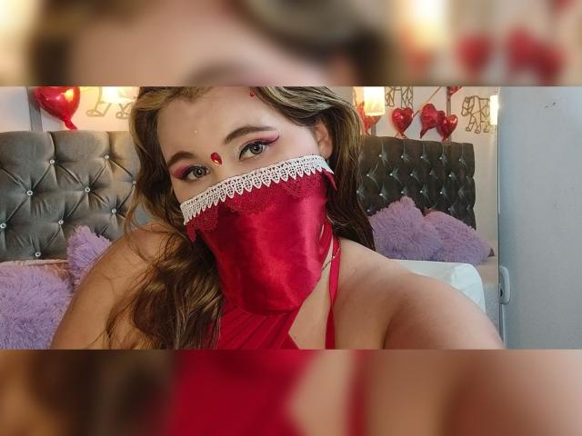 SalmaAzaman with red mask, lace trim, and red makeup dot, sitting on bed with purple pillows, wearing red lingerie, showing her tits, brown wavy hair, and pink eye makeup.