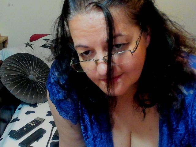 ReifeIri-hot with black hair, glasses, blue lace top, and cleavage, sitting on bed, slightly smiling, with remote control and cushion in background.