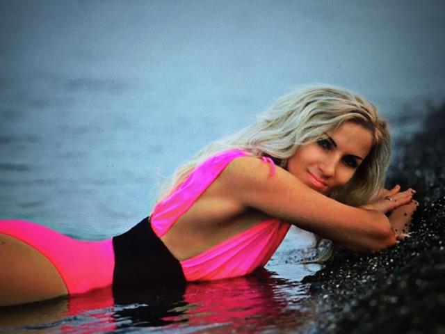 DAkotaHotti69, with long blonde hair, laying in shallow water, wearing a neon pink and black one-piece swimsuit, arms folded, looking at the camera, butt slightly raised.