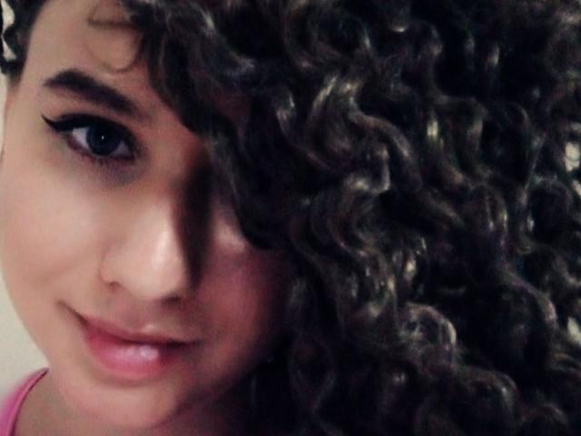 MarlanViviane has curly dark brown hair covering half her face, blue eyes with black eyeliner, and a subtle smile. She's wearing a pink shirt, showing her cute nose.