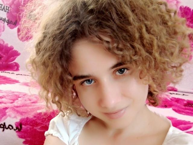 MarlanViviane has curly brown hair, blue eyes, and a subtle smile. She's wearing a white shirt, looking cute and sexy with a soft, pretty face.