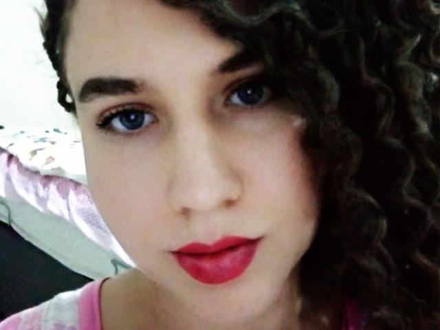 MarlanViviane has curly brown hair, blue eyes, and red lipstick. She's looking at the camera with a slight smile, wearing a pink top. Her face is centered in the image.
