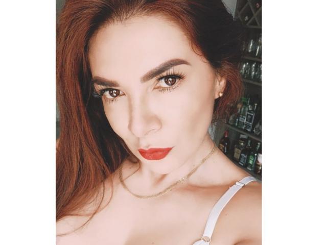 JoTaylor tem cabelo ruivo, olhos castanhos e batom vermelho, veste uma blusa branca com alças e uma corrente dourada, e olha diretamente para a câmera com uma expressão séria..
