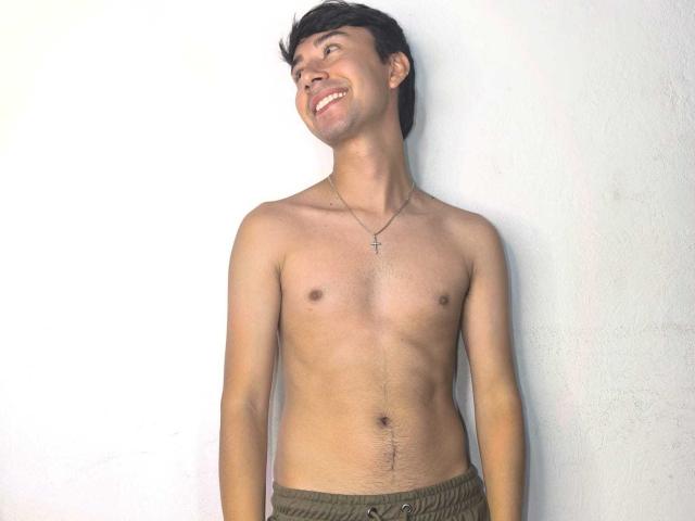 OliverHot69 is a shirtless dude with a slim build, short dark hair, and a cross necklace, smiling up at the sky, wearing green pants, and showing a belly scar.