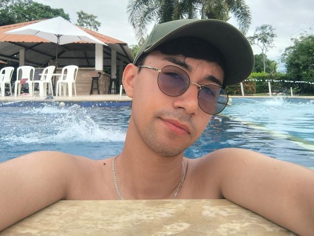 OliverHot69, com pele bronzeada, cabelo curto e escuro, boné verde, óculos de sol redondos, colar, sem camisa, apoiado na borda de uma piscina, com água espirrando, e palmeiras ao fundo..