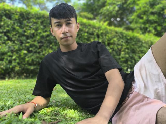 OliverHot69, with short black hair and light skin, sits on grass in a black t-shirt and pink shorts, looking slightly annoyed, right arm resting on ground.