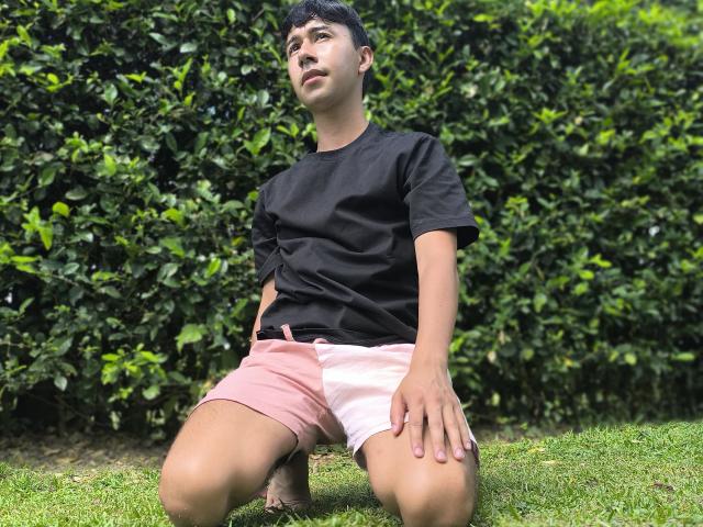 OliverHot69, à genoux sur l'herbe, portant une chemise noire et un short rose, les mains sur les cuisses, regardant vers le haut, cheveux noirs courts, peau de couleur moyenne, environnement extérieur décontracté..