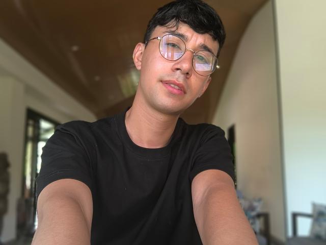 OliverHot69, avec des cheveux noirs courts, des lunettes et un t-shirt noir, souriant légèrement, autoportrait, en intérieur, bras tendus, plafond visible, décontracté, attrayant, peau de couleur moyenne, légère barbe de quelques jours..