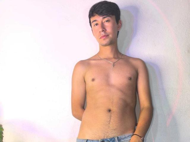 OliverHot69, shirtless, standing against a white wall, with hands behind his back, wearing blue jeans, has a slim, toned body, small nipples, and a slight belly, short black hair.