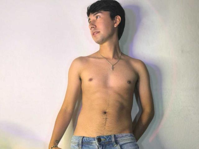 OliverHot69, torse nu avec une croix en pendentif, a une poitrine fine et poilue, et un tatouage légèrement visible sur son bras droit, il porte un jean bleu et se tient devant un mur blanc..