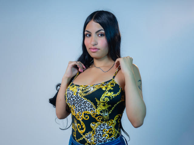 Tiffany Hils, com cabelos longos e pretos, vestindo uma regata estampada com padrão de leopardo e amarelo, segurando as alças, mostrando seus seios grandes e batom rosa, olhando para a câmera..