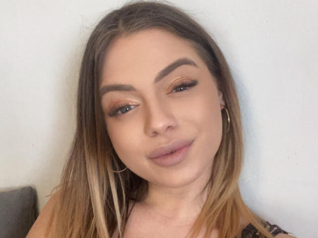 BritnyDesire has blonde hair, subtle makeup, and hoop earrings, with a slight smile and half-closed eyes. She's looking at the camera with a relaxed, cute expression.