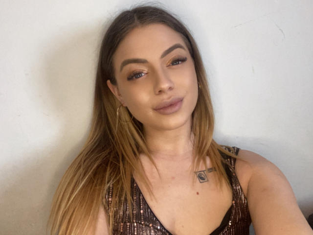 BritnyDesire has long brown hair, a small tattoo near her collarbone, and is wearing a shiny brown dress with a low neckline, showing her small tits. She's smiling slightly.